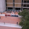 Aerial view of the University of Arizona campus plaza featuring the large red and blue “BEAR DOWN” sign, with students walking, biking, and sitting at tables outside a modern campus building.