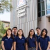 A group of seven University of Arizona Global Ambassadors stand together outside the Global Center, smiling at the camera. They are wearing matching navy polo shirts with the Arizona International logo, with the building’s large "A" logo and "Global Center" signage visible behind them.