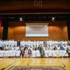 Members of the University of Arizona Gulf Cooperation Council (GCC-UA) alumni community pose for a large group photo on stage during the 11th Annual GCC Alumni Reunion in Salalah, Oman. The audience includes alumni, officials, and guests seated and standing in several rows inside an auditorium decorated with red curtains and projection screens.