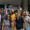 A group of seven students wearing matching orange sunglasses smile and pose together at an indoor event. They make hand gestures, including the University of Arizona "Wildcat" hand sign, while enjoying the social atmosphere.