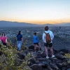 Four people stand on rocky terrain at sunset, overlooking the city of Tucson, Arizona. The view includes the sprawling cityscape below with distant mountains and a colorful sky.