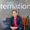 Hassan Hijazi, Senior Assistant Vice President for University of Arizona International, sits in a chair in the Arizona International office. He is wearing a navy blue suit with a red shirt, smiling at the camera. Behind him, the word “International” is displayed prominently on the wall.