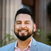 Portrait of Ricky Salazar smiling outdoors in front of a building with columns. He is wearing a light blue blazer and a pink collared shirt.