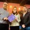 Three people stand together indoors under warm hanging lights, smiling at the camera. The person on the left holds a purple and blue booklet. The background features a chalkboard-style mural with text and illustrations, giving the space a casual, creative atmosphere.