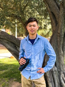 Dennis stands in front of a tree holding a small video camera