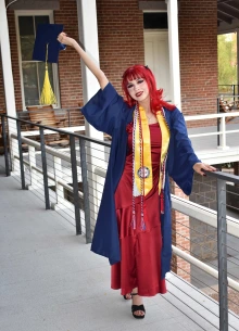 A joyful graduate with bright red hair poses on a covered outdoor walkway in front of a brick building. She wears a deep red dress beneath a navy blue graduation gown and holds a matching cap with a gold tassel raised high in one hand. She also wears a gold stole and multiple honor cords, smiling confidently at the camera. The setting suggests a celebratory moment on the University of Arizona campus.