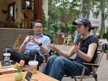 A man and woman sit outdoors at a café table, engaged in conversation. The man, wearing glasses and a light blue dress shirt, listens attentively with his hands clasped. The woman, wearing a dark t-shirt and baseball cap, gestures with her hands while speaking. Drinks with straws sit on the table in front of them.