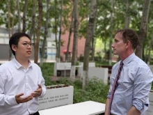 Two men stand outdoors in conversation, surrounded by tall trees and greenery. The man on the left, wearing glasses and a white button-up shirt, gestures with his hands while speaking. The man on the right, wearing a striped tie and rolled-up shirt sleeves, listens attentively.