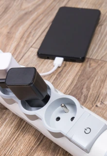 A smartphone charging through a USB cable connected to a power strip with multiple plug adapters on a wooden floor.