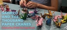 Person's hands shown making origami paper cranes with 20 colorful cranes on table 
