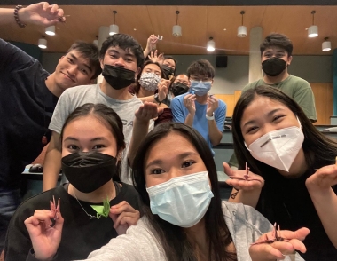 Nine students, some wearing face masks, posing for the camera holding origami paper cranes they have made