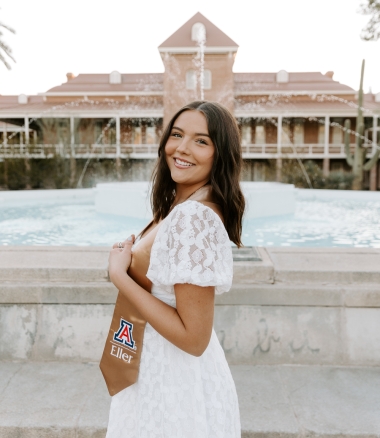 Madison in white dress in front of Old Main Fountain, wearing a gold Eller graduation stole with Block A logo