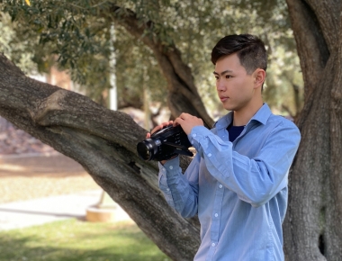 Dennis Zhuang with camera in front of a tree on campus