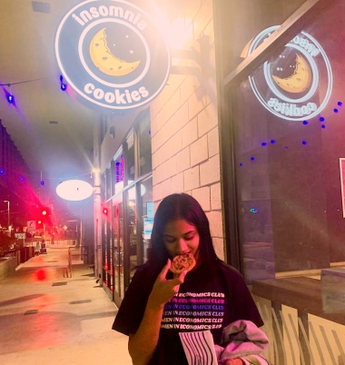 Ananya is standing under a sign for Insomnia Cookies, eating a cookie, and wearing a t-shirt that says Women in Economics Club repeated six times in several colors. 