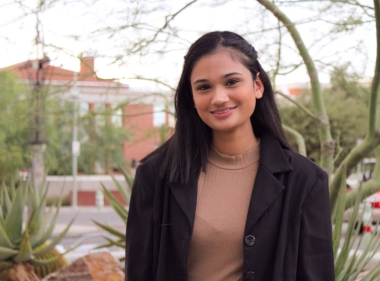 Ananya is standing in front of a palo verde tree on the UArizona campus. She is wearing a tan sweater and a black suit jacket.