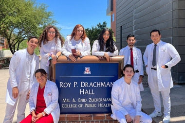 Eight UVM students wearing white medical coats are standing surrounding the MEXCOPH building sign